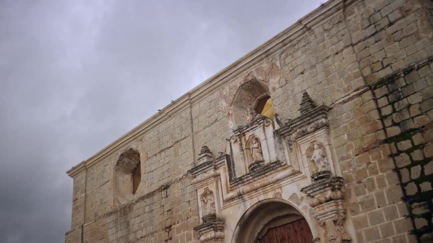 A lowangle shot shows the weathered stone exterior of an old colonial church under a grey overcast sky highlighting the arched wooden entrance and intricate religious carvings set into the masonry of the building facade