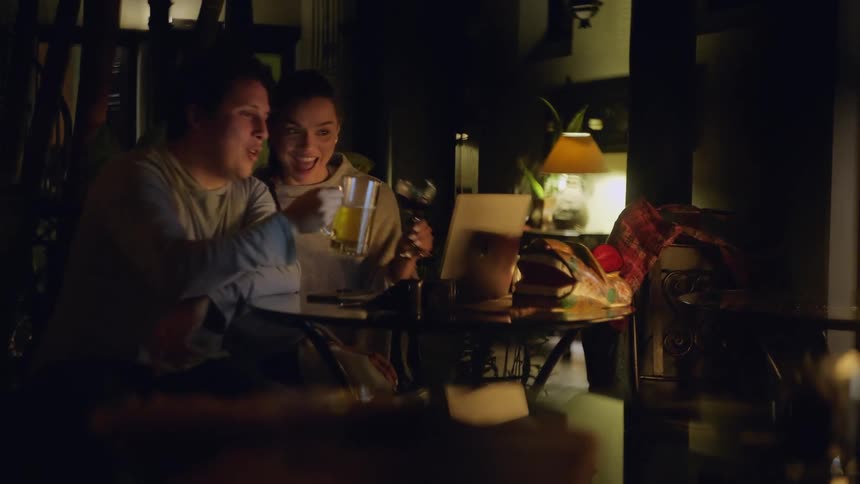 A man and a woman sit at a dark oval table laughing and talking while looking at a laptop screen and then they raise a beer mug and a wine glass to toast toward the camera during a video call surrounded by warm ambient lighting