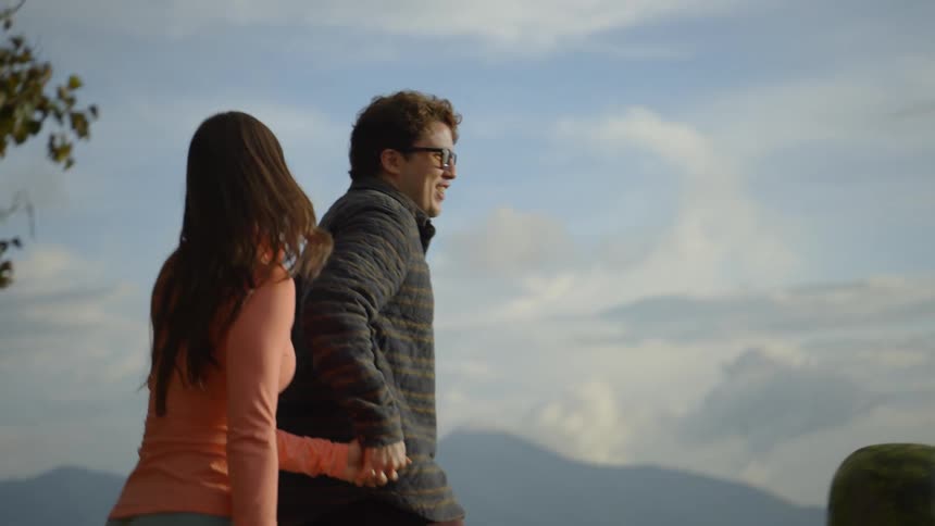A young couple holding hands in excitement walking toward the statue palm at hobbitenango ecopark at midday the man smiling behind his glasses while the woman in an orange shirt looks out at the surrounding nature