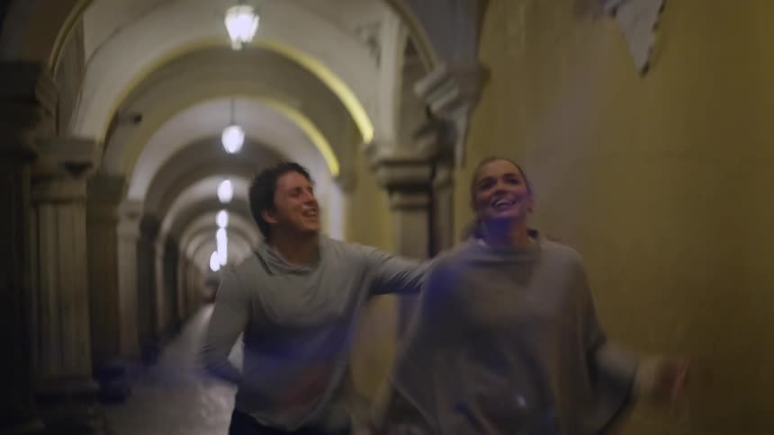 A man and woman run through a dim arched stone corridor their motion blurred as they move forward