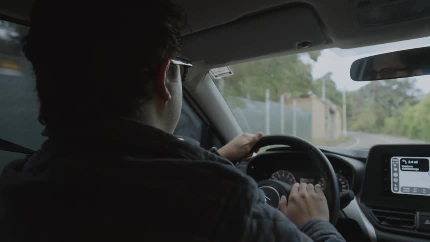 A man driving on a countryside road at midday wearing sunglasses and tapping on the steering wheel as he drives