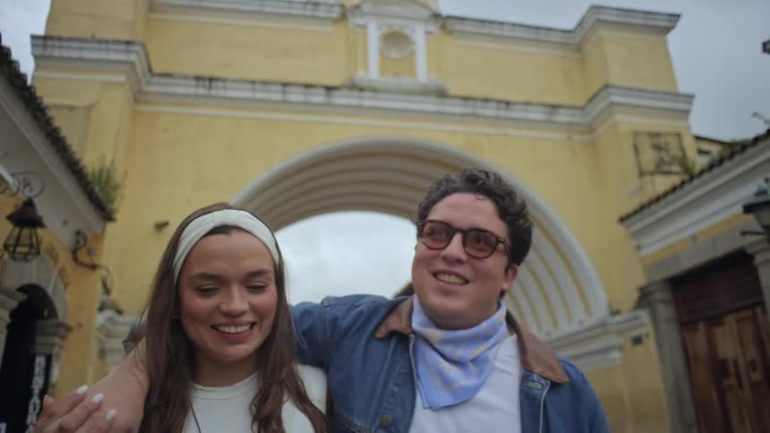 A smiling couple walks arminarm past a grand yellow archway in a historic town engaged in a cheerful conversation while enjoying their morning stroll through the scenic urban landscape