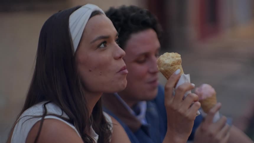 Two people seated outdoors each holding and eating an ice cream cone with their hands and faces in partial profile against a softly blurred street background