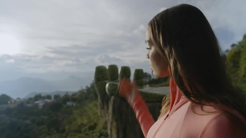 A woman wearing a pink zipup athletic top stands at a scenic mountain overlook lifting her hand to shield her eyes from the sunlight as she gazes thoughtfully across a lush green valley and distant hills under a bright hazy sky