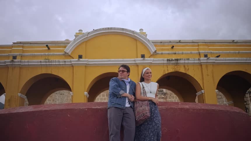 A man with glasses and a denim jacket stands next to a woman wearing a white headband and a blue patterned skirt as they pose in front of a large yellow arched building under an overcast sky