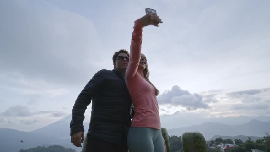 Couple outdoors taking a selfie with a smartphone with mountains and a cloudy sky in the background
