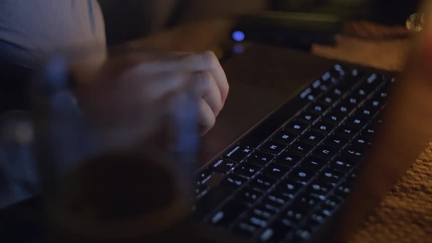 A person uses a laptop trackpad in a dimly lit indoor setting with the backlit keyboard glowing in the darka glass containing a dark liquid sits in the foreground partially blurring the view of the persons hand as they navigate the computer