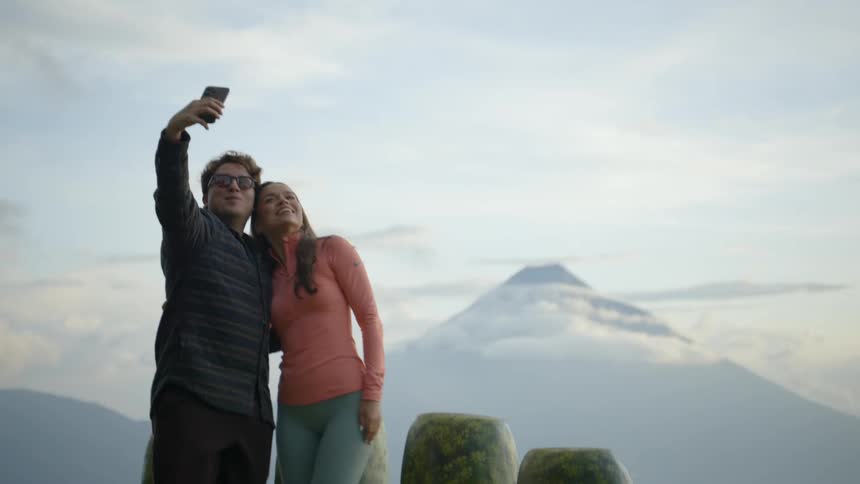Two people standing close together on a scenic overlook with one holding a smartphone raised to take a photo while the other leans in and a large conical mountain rising in the distance above low clouds