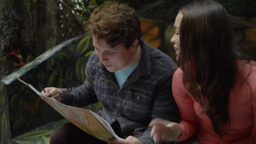 A young man and woman sit together on a bench outdoors intently studying a large paper map to find their way