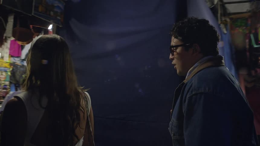 A man in a denim jacket and a woman with a white headband walk through a cramped dimly lit night market filled with hanging garments and various merchandise