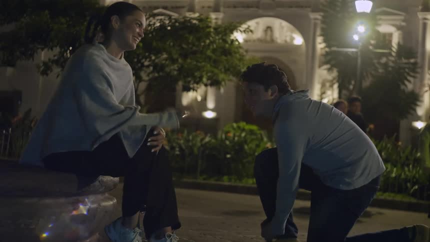 A woman sits on a stone ledge at night while a man kneels to tie her shoelace in front of a historical building