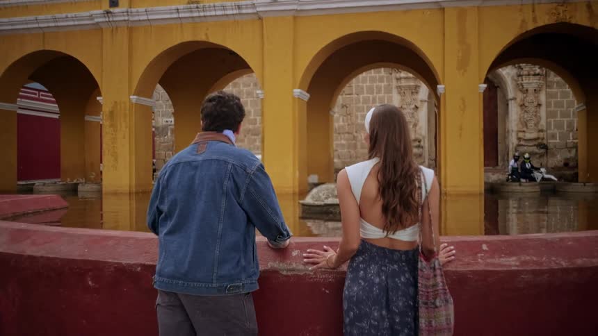 A young couple walks towards and then leans on a red wall looking at a large historic yellow fountain with prominent archways
