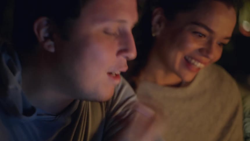 The video features two people sitting together in a dimly lit environment their faces illuminated by a warm glow from belowthe man in the foreground laughs and eats while looking at something offcamera as the woman beside him watches with a bright