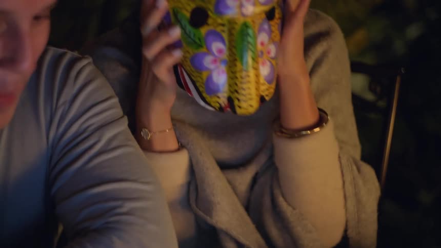 A woman examines a brightly decorated traditional mask with a radiant smile before pressing it against her face to playfully surprise the man sitting beside her as warm evening light illuminates the cozy scene