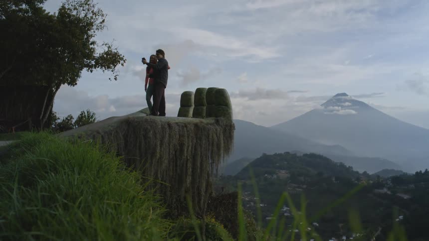 A man and woman take a selfie on a giant stone hand sculpture overlooking a valley and a volcano before walking back toward a grassy path during a cloudy day