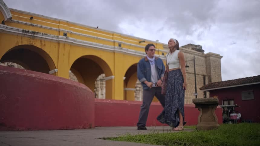 A young couple walking into a public area and sitting side by side on a bench the man wearing sunglasses and a jean jacket and the woman carrying a handbag with a yellow building in front of them