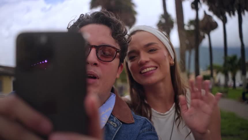 A man with glasses and a woman in a headband smile and wave during a video call on a smartphone while standing in a town square with palm trees and buildings