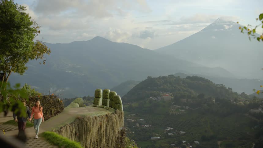 A man and woman stand on a stone platform shaped like a giant hand while taking a selfie against a backdrop of mountains they then walk away from the scenic overlook leaving the handshaped structure empty as it faces the valley below