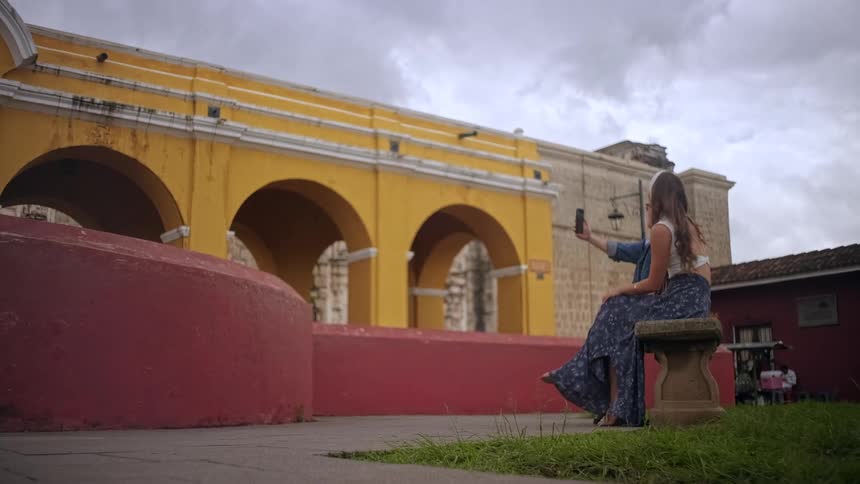 A happy couple takes a selfie while sitting on a stone bench in a historic plaza under a cloudy sky