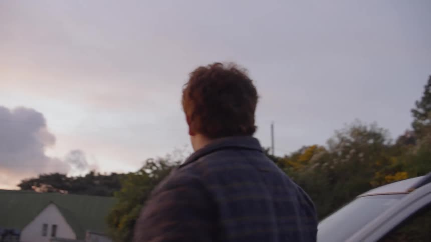 A man in a flannel shirt and a woman in a poncho exit a white car at dusk then walk toward a scenic overlook where the man puts his arm around her as they look at the clouds