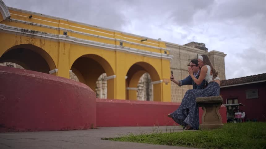 Two people sit on a stone bench in an open plaza with one holding a phone outward for a selfie while a yellow arched building red walls grass and a cloudy sky form the architectural backdrop
