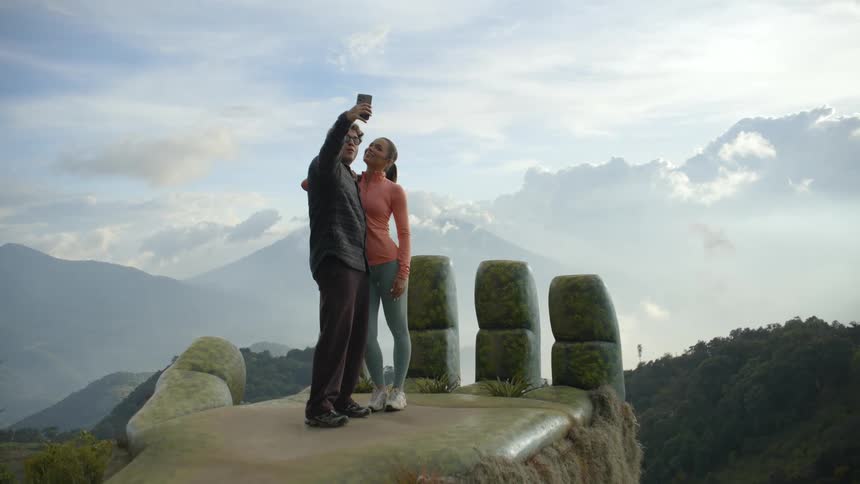 Couple standing at an outdoor viewpoint taking a selfie with a smartphone with mountains and a clear sky in the background