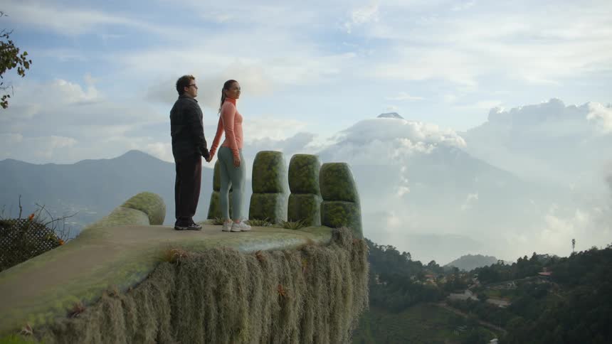 A man and woman walk onto a large stone hand sculpture overlooking a misty mountain range where they hold hands and look out at the horizon together