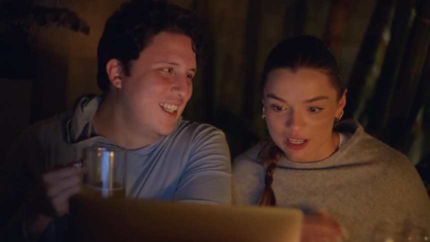 Couple seated at a table looks at an illuminated screen while holding a drink in an indoor nighttime setting
