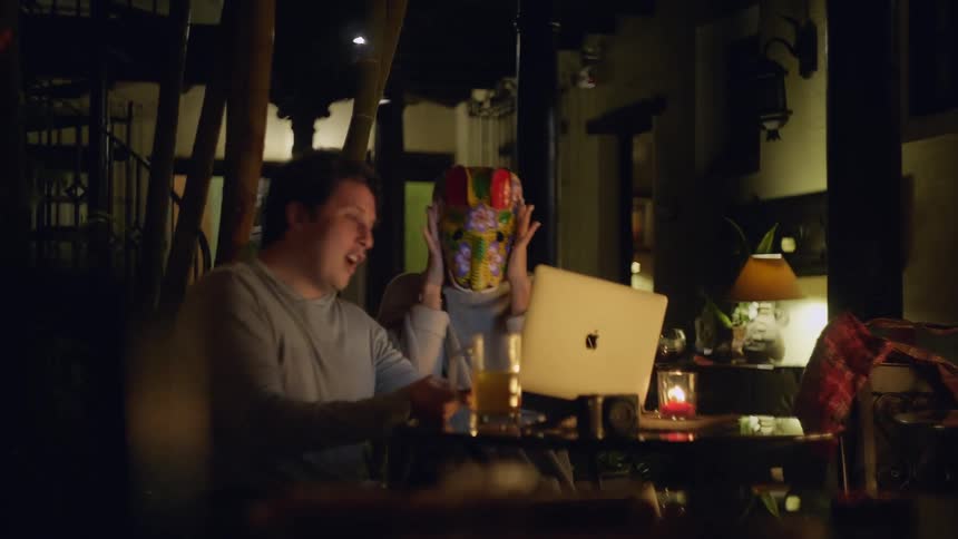 In the video a man and a woman sit at a dimly lit outdoor table laughing and interacting with someone through a laptop screen the woman holds up a colorful traditional mask to her face to playfully surprise the person on the video call