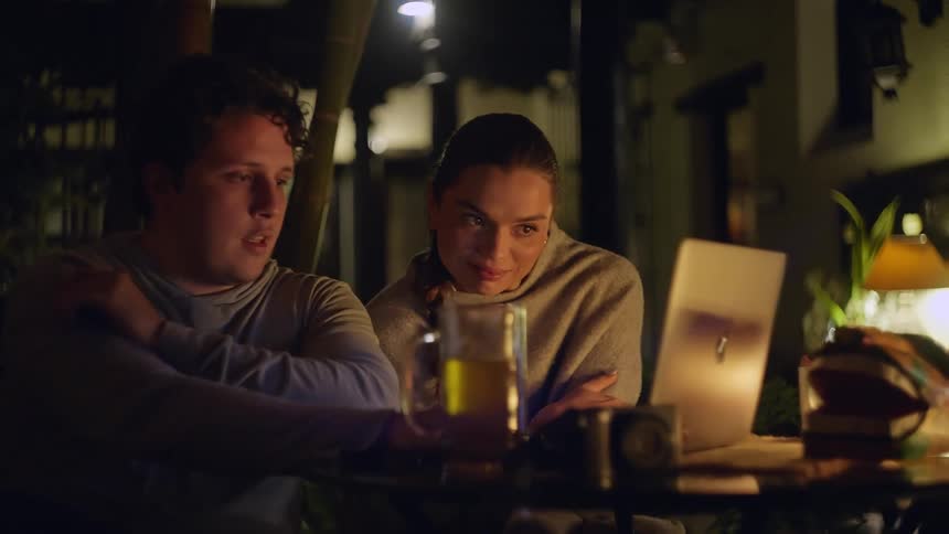 A couple sitting side by side at night the man drinking a glass of beer and the woman a glass of wine discussing something as he scrolls through a laptop in front of them