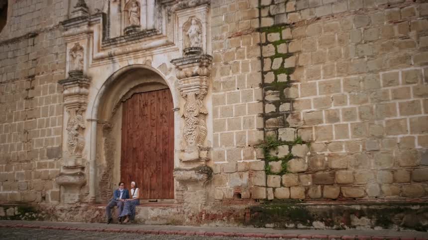 A couple sitting at the entrance of a church at midday with large brown wooden doors closed behind them as they talk