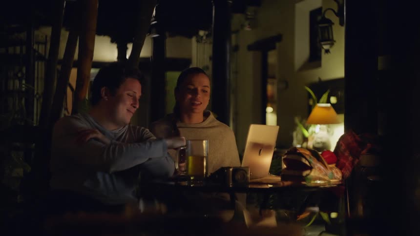 Couple seated at an outdoor table at night using a laptop with drinks and personal items on the table