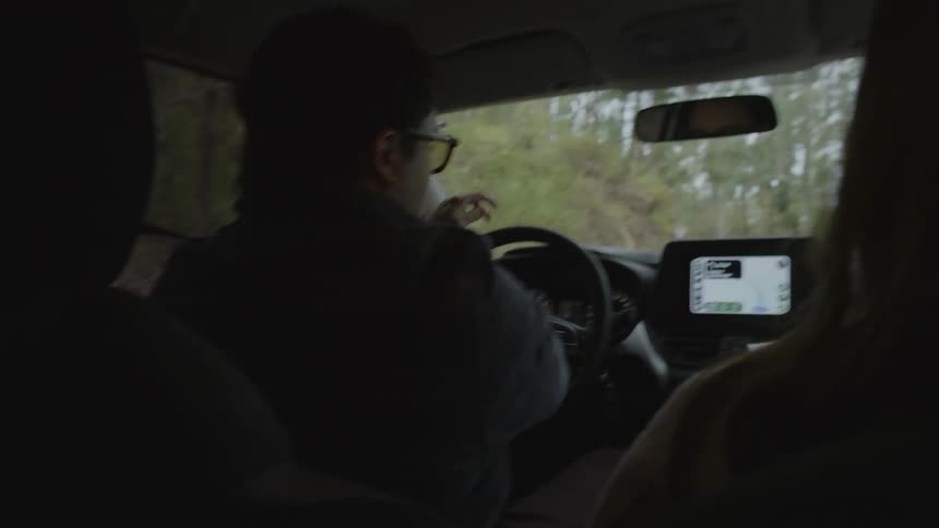 A couple driving through the wooded countryside at midday the man wearing sunglasses as other cars pass by