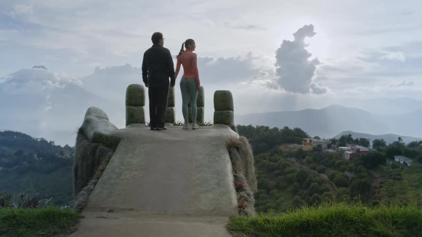 A couple stands on a stone overlooking across a lush mountain valley toward distant hills