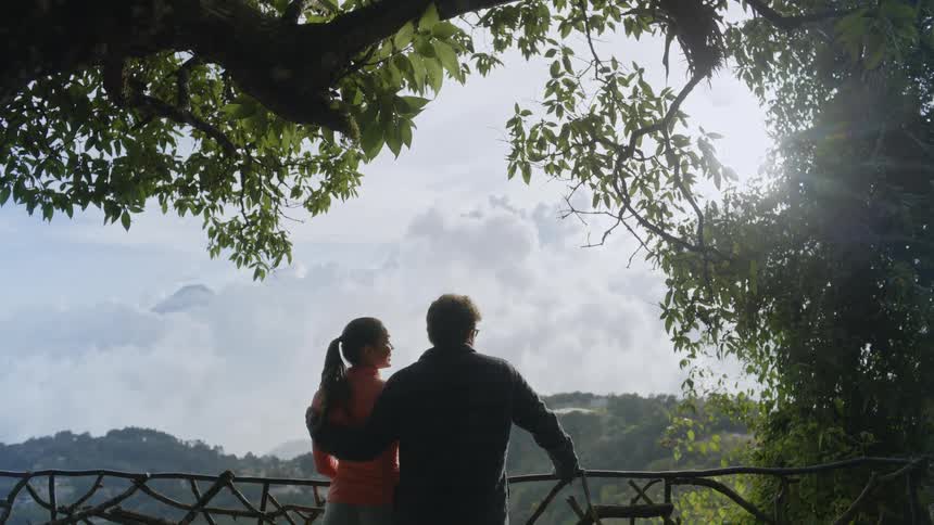 A couple stands at a scenic overlook framed by lush green trees looking out over a vast valley filled with clouds other hikers occasionally walk past them as they admire the bright misty mountain landscape from behind a wooden railing