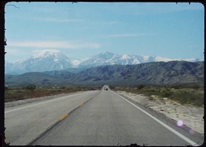 A long straight road stretches toward a horizon of majestic snowcapped mountains under a clear blue sky the video features a vintage grainy film aesthetic that captures the vastness of a desert landscape during a road trip