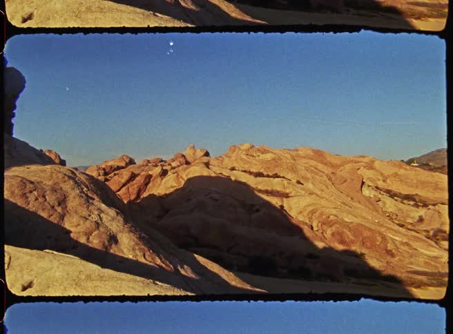 A wide shot reveals a vast desert landscape featuring jagged sandstone rock formations under a clear blue sky during the golden hour with long shadows stretching across the arid terrain and distant hills visible on the horizon