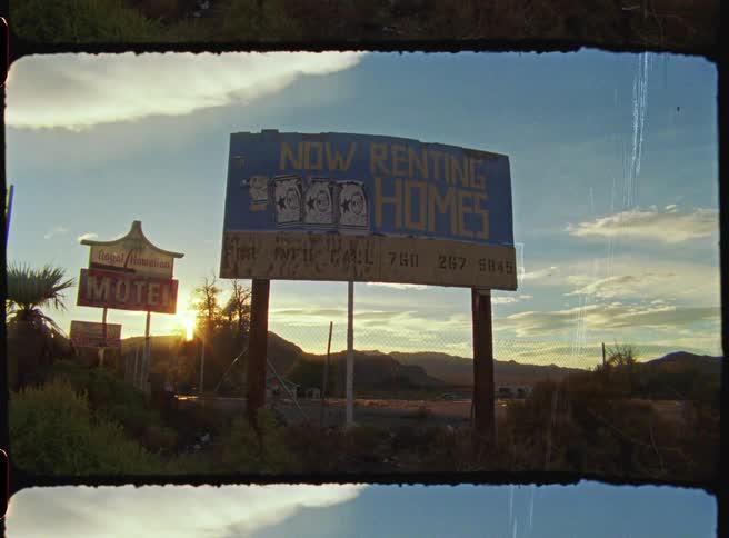 A giant rent billboard and a motel sign standing in a desert landscape during sunset as light flares behind the mountains