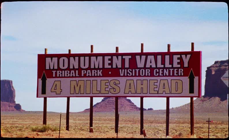 A vintagestyle sign for monument valley stands alongside a desert road pointing toward the towering sandstone buttes that rise from the red earth under a clear blue sky as a vehicle passes by