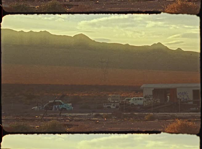 A bird flies across a desolate desert landscape featuring a graffiticovered car and an abandoned shack all silhouetted against rugged mountains during the warm glow of a golden sunset