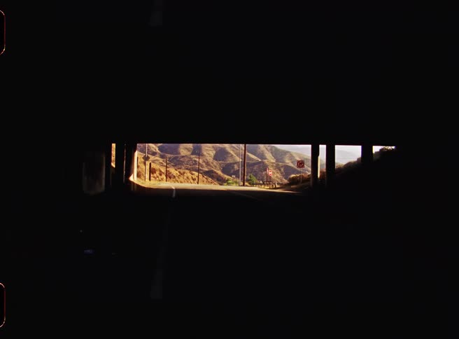 Road seen through a dark underpass with a sunlit mountainous landscape in the distance