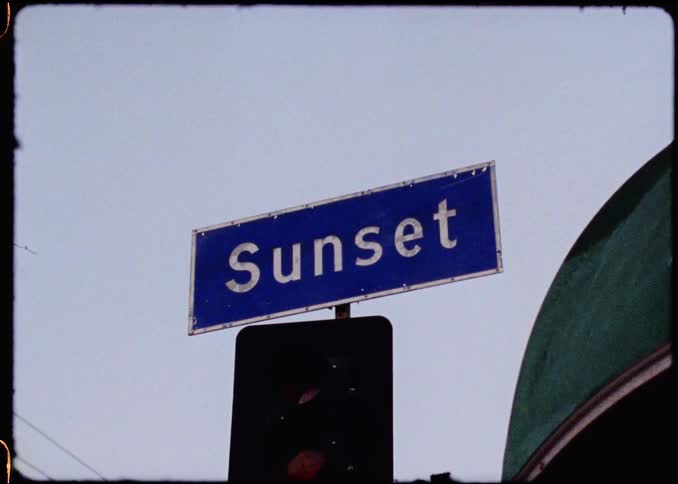 A close shot shows a blue street sign with sunset written in white text positioned above a traffic light