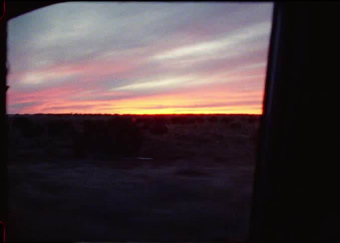A vibrant pink and orange sunset over a dark landscape is viewed from the window of a moving car