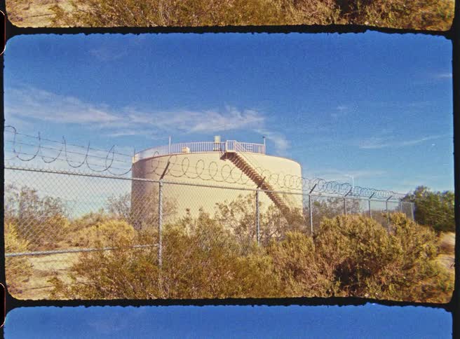 A large cylindrical water tank stands behind a barbed wire security fence in a dry desert landscape under a clear blue sky featuring a metal staircase leading to the top of the structure surrounded by arid vegetation and scrubland