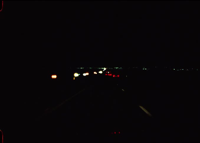 The video shows a pointofview shot from a vehicle driving down a dark highway at night glowing headlights and taillights from distant traffic create small spots of color against the deep black surroundings