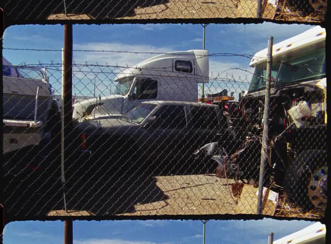 A garage filled with junk cars and trucks enclosed by a wiremesh fence on a sunny day
