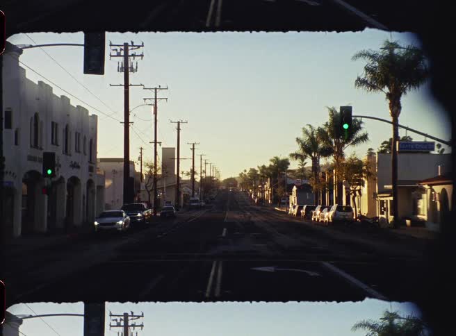 A long straight road stretches toward the horizon during a golden hour sunset flanked by utility poles and palm treesparked cars and lowrise buildings line the quiet street under a clear sky as a green traffic light glows in the distance
