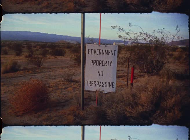 A white sign standing in a dry desert landscape warns that the area is government property and prohibits trespassing the footage is shot in a vintage film style showing scrub brush and distant mountains under a pale sky