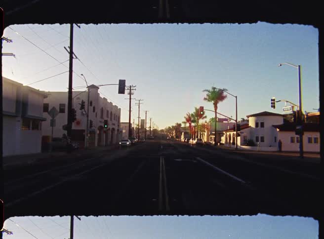 An empty city street lined with buildings and palm trees filmed with a vintage aesthetic under a bright lowhanging sun