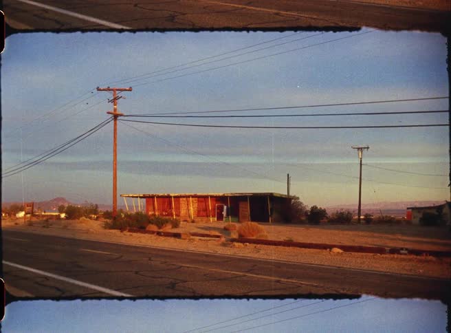 This vintage filmstyle footage depicts a weathered singlestory building situated in a vast desert landscape alongside a paved road utility poles and power lines stretch across the scene under a clear sky during the warm light of golden hour